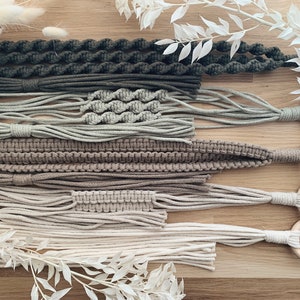 Könnte beinhalten: Ein Set aus fünf Makramee-Pflanzenhängern in verschiedenen Brauntönen, Beige und Grün. Die Hänger sind aus dickem Baumwollgarn gefertigt und weisen unterschiedliche Knotenmuster auf. Jeder Hänger ist an einem Holzring befestigt.