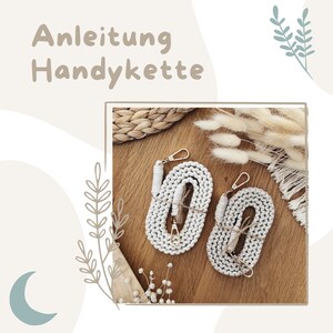Könnte beinhalten: Zwei weiße Makramee-Handyketten mit goldfarbenen Verschlüssen und einer braunen Schnur, die auf einer Holzoberfläche präsentiert werden. Der Text "Anleitung Handyette" befindet sich oben im Bild.