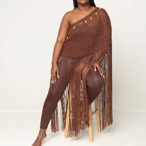 May include: A brown crochet top with fringe detailing and gold accents. The top is one-shoulder and has a loose fit. The model is wearing brown leggings.