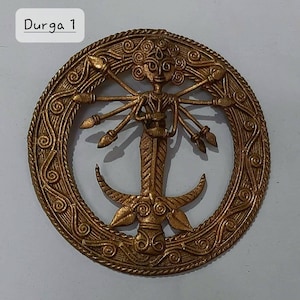 Puede incluir: Un colgante de metal dorado que representa a la diosa hind&uacute; Durga. El colgante presenta un dise&ntilde;o circular con detalles intrincados y una representaci&oacute;n de Durga con m&uacute;ltiples brazos que sostienen armas. El texto "Durga 1" es visible en la parte superior de la imagen.