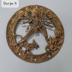 Puede incluir: Un colgante de pared circular de lat&oacute;n que representa a la diosa hind&uacute; Durga, montando un le&oacute;n y matando a un demonio. El dise&ntilde;o intrincado presenta tallas detalladas y un acabado dorado.