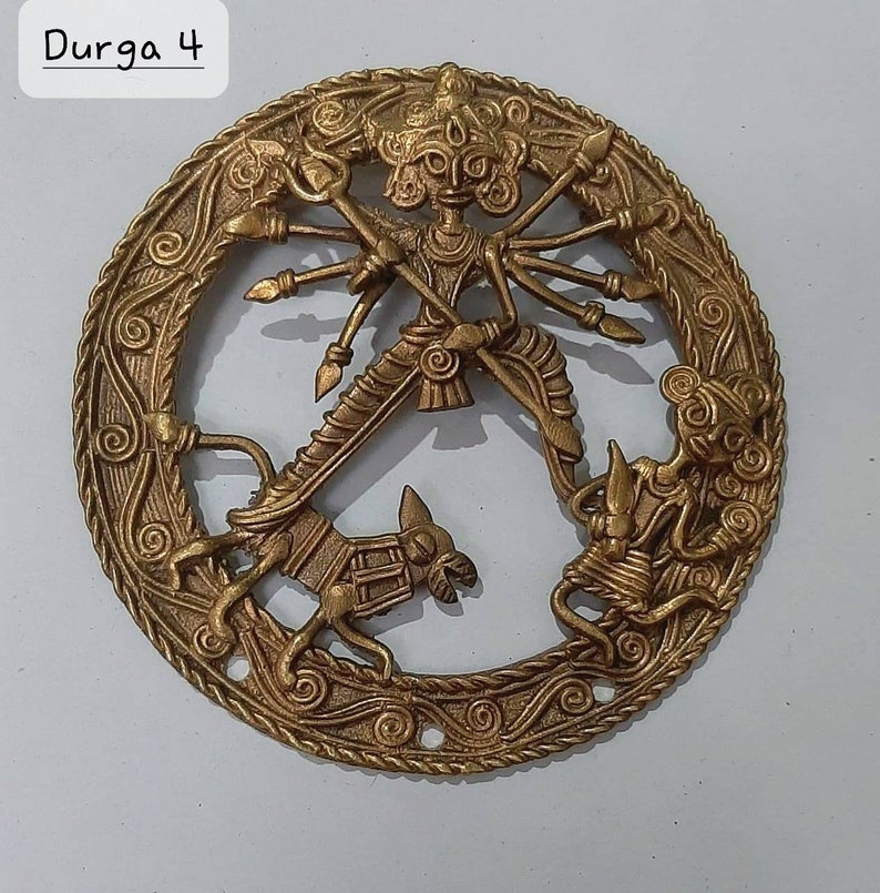 Puede incluir: Un colgante de pared circular de lat&oacute;n que representa a la diosa hind&uacute; Durga, montando un le&oacute;n y matando a un demonio. El dise&ntilde;o intrincado presenta tallas detalladas y un acabado dorado.