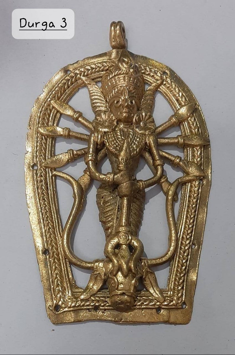 Puede incluir: Un colgante de metal dorado que representa a la diosa hind&uacute; Durga. El colgante tiene forma de herradura y presenta un dise&ntilde;o detallado de Durga con m&uacute;ltiples brazos que sostienen armas. El colgante est&aacute; inscrito con el texto "Durga 3".