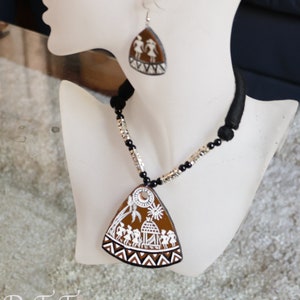 May include: A brown and white ceramic necklace with a triangular pendant featuring a painted scene of people dancing around a house. The pendant is attached to a black and silver beaded chain. The necklace is displayed on a white mannequin.