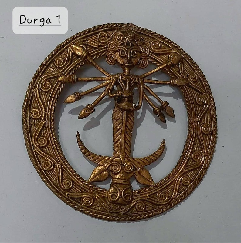 Puede incluir: Un colgante de metal dorado que representa a la diosa hind&uacute; Durga. El colgante presenta un dise&ntilde;o circular con detalles intrincados y una representaci&oacute;n de Durga con m&uacute;ltiples brazos que sostienen armas. El texto "Durga 1" es visible en la parte superior de la imagen.