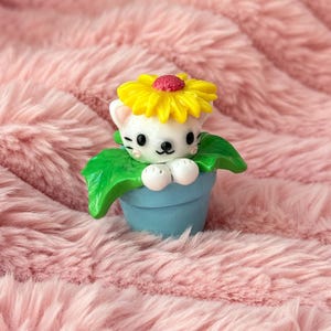 Può includere: Una statuetta stravagante di un gatto bianco con un fiore giallo in testa, che emerge da un vaso blu con foglie verdi. Il gatto ha occhi e baffi neri. La statuetta è ambientata su uno sfondo rosa e soffice.