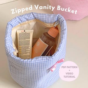 Może przedstawiać: Niebiesko-biały, zapinany na zamek kubełek kosmetyczny w kratkę z beżowym wnętrzem, zawierający kosmetyki. Różowa kokarda jest przymocowana z boku. Tekst "Zipped Vanity Bucket" znajduje się na górze. Różowy kubełek w kratkę jest w tle. Tekst po prawej stronie: "PDF Pattern + Video Tutorial".
