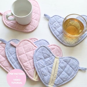 Puede incluir: Posavasos de tela acolchada en forma de corazón con estampados de cuadros rosas y azules. Los posavasos tienen un lazo para colgar y se muestran con una taza blanca y un vaso de té. La imagen también incluye el texto "PDF Pattern + Video Tutorial".