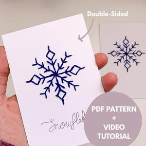 May include: A white card featuring a blue snowflake design. The card is double-sided, with the snowflake on both sides. The word "Snowflake" is written on the card. A circular graphic states "PDF Pattern + Video Tutorial".