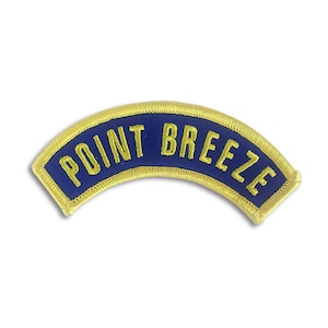 Pode incluir: Um remendo bordado azul e dourado com o texto "POINT BREEZE" em forma curva.