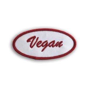 Pode incluir: Etiqueta oval branca com uma borda vermelha bordada. A palavra "Vegan" está escrita em letras cursivas vermelhas.