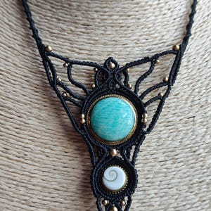 Pode incluir: Colar de macramé preto feito à mão com um pingente grande e redondo de pedra verde-azulado claro com uma moldura dourada. O colar também inclui um pingente branco menor com um design em espiral e contas douradas.