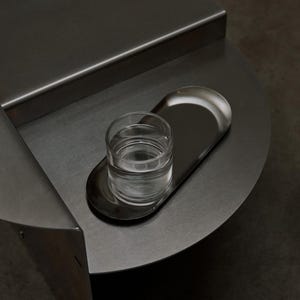 May include: A dark gray metal side table with a circular top and a raised back edge. An oval-shaped metal tray sits on the surface, holding a clear glass tumbler filled with water. The table features a smooth, matte finish and a geometric design.