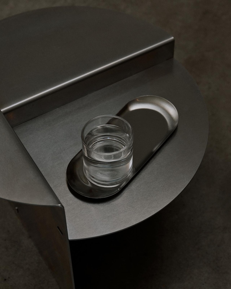 May include: A dark gray metal side table with a circular top and a raised back edge. An oval-shaped metal tray sits on the surface, holding a clear glass tumbler filled with water. The table features a smooth, matte finish and a geometric design.