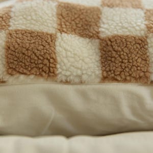 May include: A close-up of a brown and white checkered throw pillow with a soft, fuzzy texture. The pillow is made of a plush fabric and has a subtle sheen.