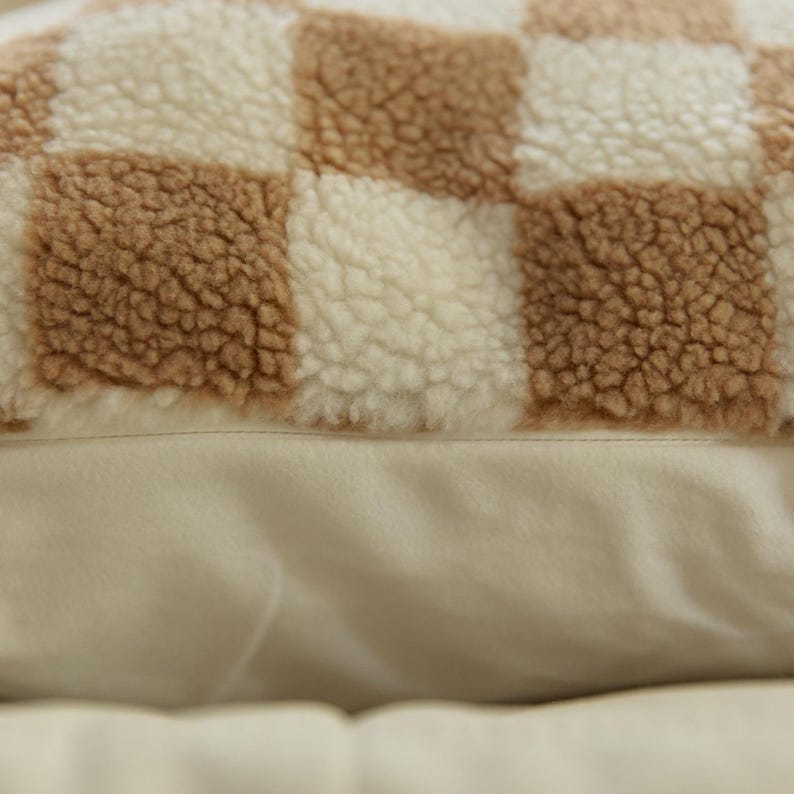 May include: A close-up of a brown and white checkered throw pillow with a soft, fuzzy texture. The pillow is made of a plush fabric and has a subtle sheen.