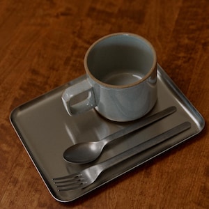 May include: A light blue ceramic mug with a handle sits on a rectangular metal tray, accompanied by a spoon and fork. The mug has a textured surface and a neutral color. The spoon and fork have a matte finish.