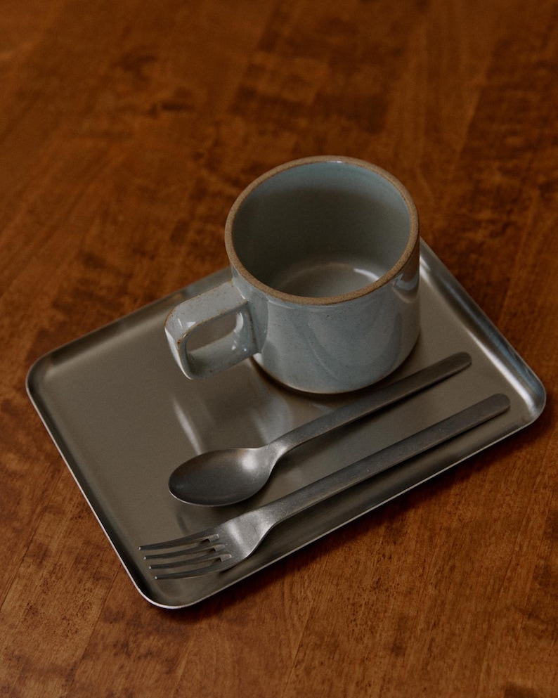 May include: A light blue ceramic mug with a handle sits on a rectangular metal tray, accompanied by a spoon and fork. The mug has a textured surface and a neutral color. The spoon and fork have a matte finish.