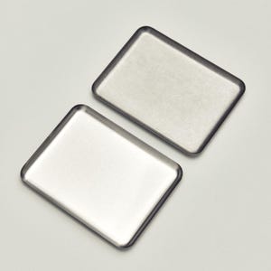 May include: Two rectangular, stainless steel trays with rounded corners are shown against a white backdrop. The trays have a brushed metal finish, reflecting the light. The trays are likely used for serving or organizing small items.