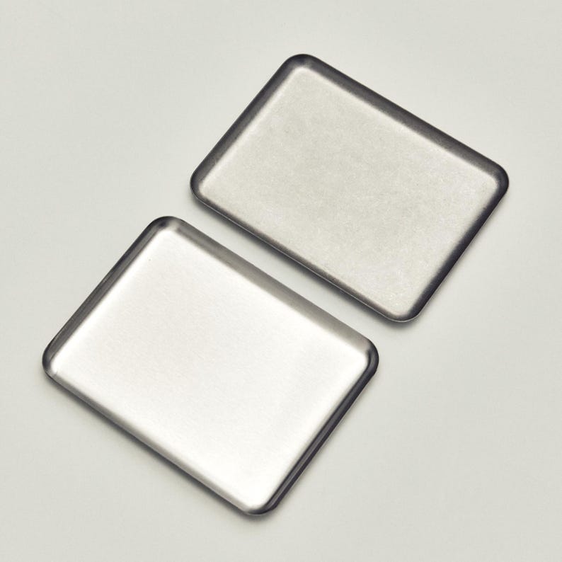 May include: Two rectangular, stainless steel trays with rounded corners are displayed against a white background. The trays have a brushed metal finish, reflecting the light. The trays are likely used for serving or organizing small items.