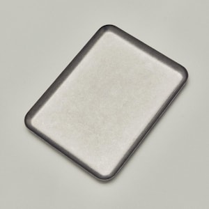 May include: A rectangular, silver-toned tray with a dark gray rim. The tray has a slightly textured surface and rounded corners, set against a neutral background. The tray's design is simple and modern.