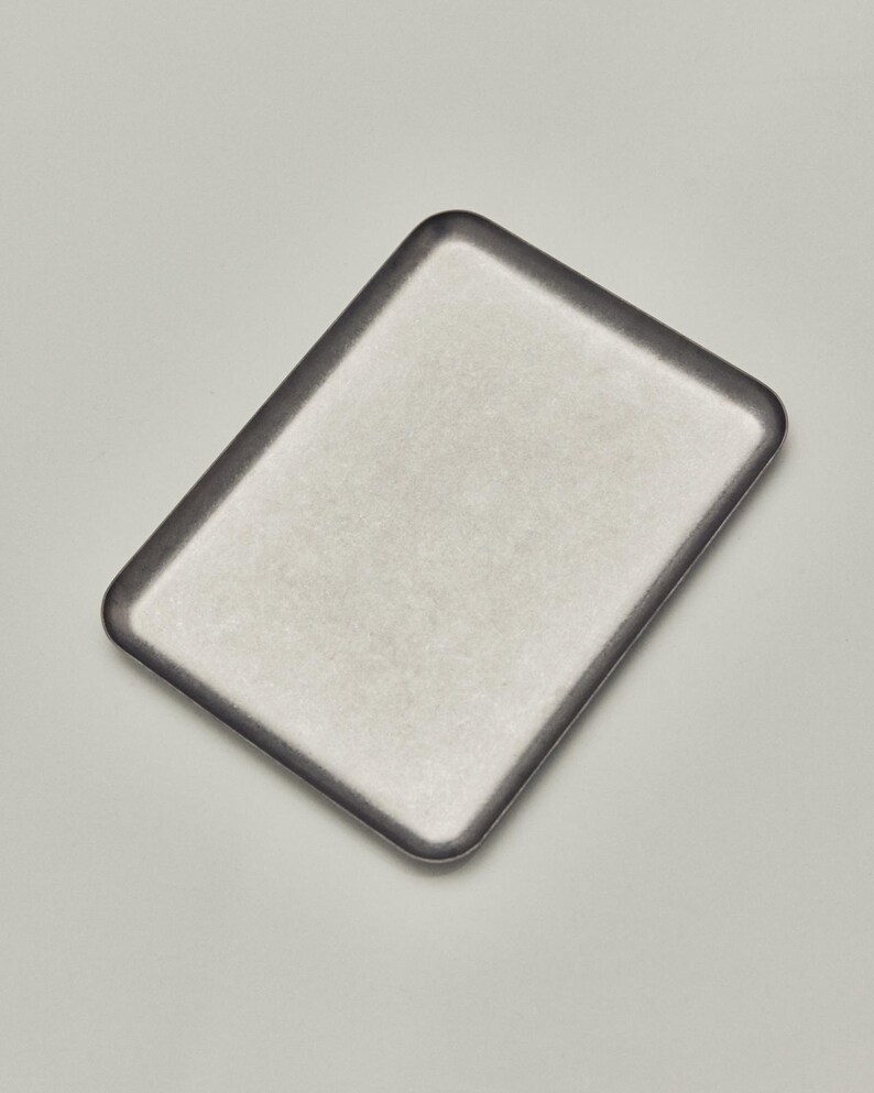 May include: A rectangular, silver-toned tray with a dark gray rim. The tray has a slightly textured surface and rounded corners, set against a neutral background. The tray's design is simple and modern.