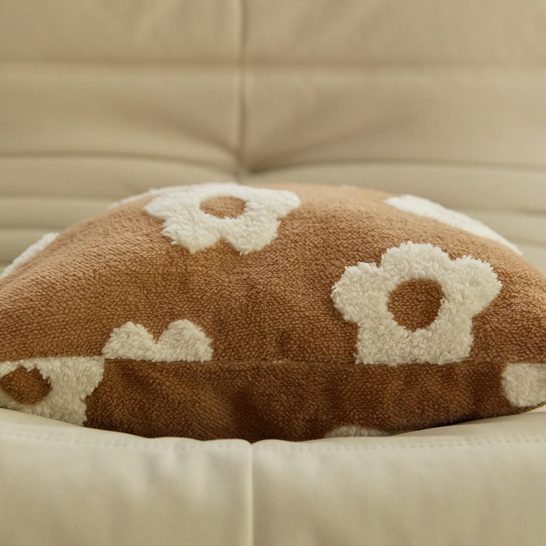 May include: A brown throw pillow with white fluffy daisy-shaped designs. The pillow is made of a soft, plush fabric.