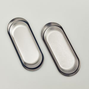 May include: Two oval-shaped metal trays with a smooth, brushed silver finish. Each tray features a recessed center and a raised, rounded rim, presenting a clean, uniform appearance with a metallic sheen that reflects light across the surface.