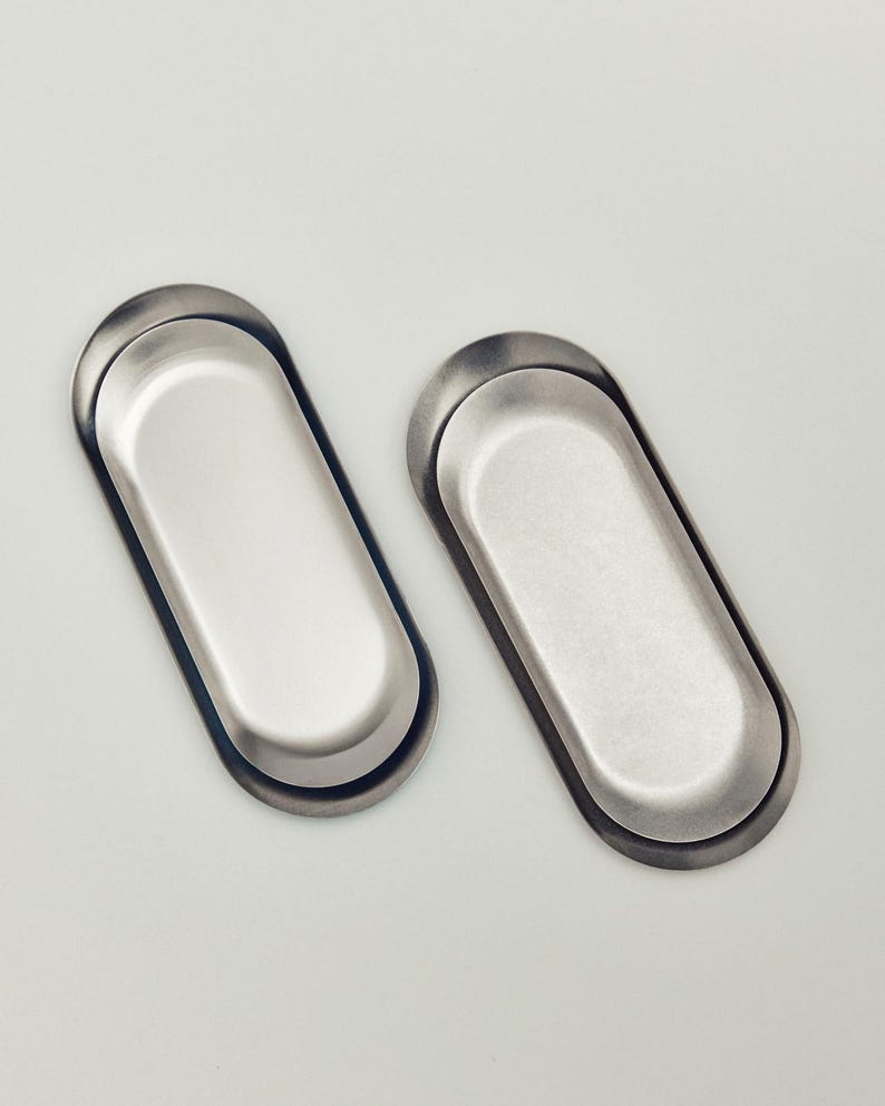 May include: Two oval-shaped metal trays with a smooth, brushed silver finish. Each tray features a recessed center and a raised, rounded rim, presenting a clean, uniform appearance with a metallic sheen that reflects light across the surface.