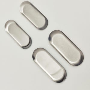 May include: Four small, elongated, oval-shaped metal trays with a smooth, brushed silver finish. Each tray features a shallow, recessed center and rounded edges, arranged in a scattered pattern across the frame, showcasing a uniform metallic texture.