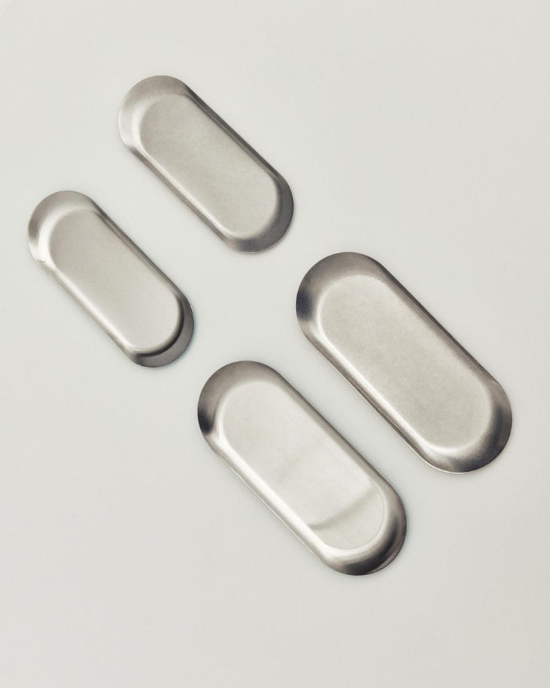 May include: Four small, elongated, oval-shaped metal trays with a smooth, brushed silver finish. Each tray features a shallow, recessed center and rounded edges, arranged in a scattered pattern across the frame, showcasing a uniform metallic texture.