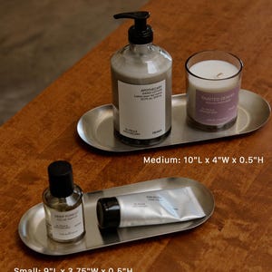May include: Two oval stainless steel trays. The medium tray, 10"L x 4"W x 0.5"H, holds a pump bottle of hand lotion and a candle labeled Painted Desert. The small tray, 9"L x 3.75"W x 0.5"H, holds a glass bottle of perfume and a tube of hand cream.