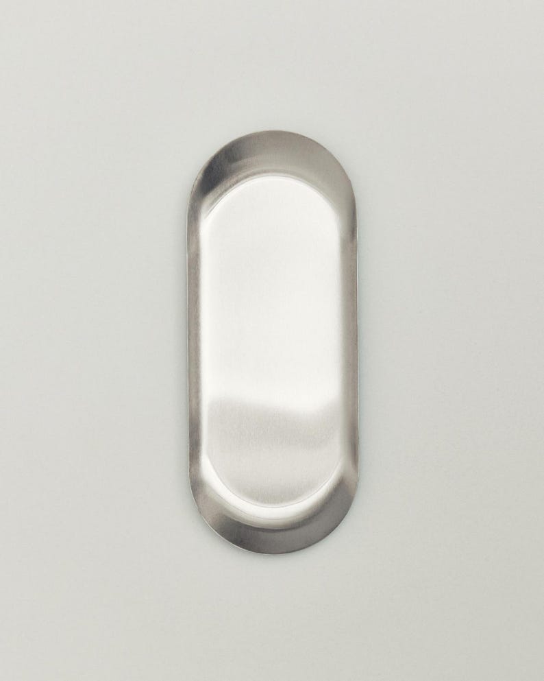 May include: A small, oval-shaped metal tray with a smooth, brushed silver finish. The tray features a shallow, recessed center and a slightly raised, rounded rim that follows the elongated oval contour of the overall object design.