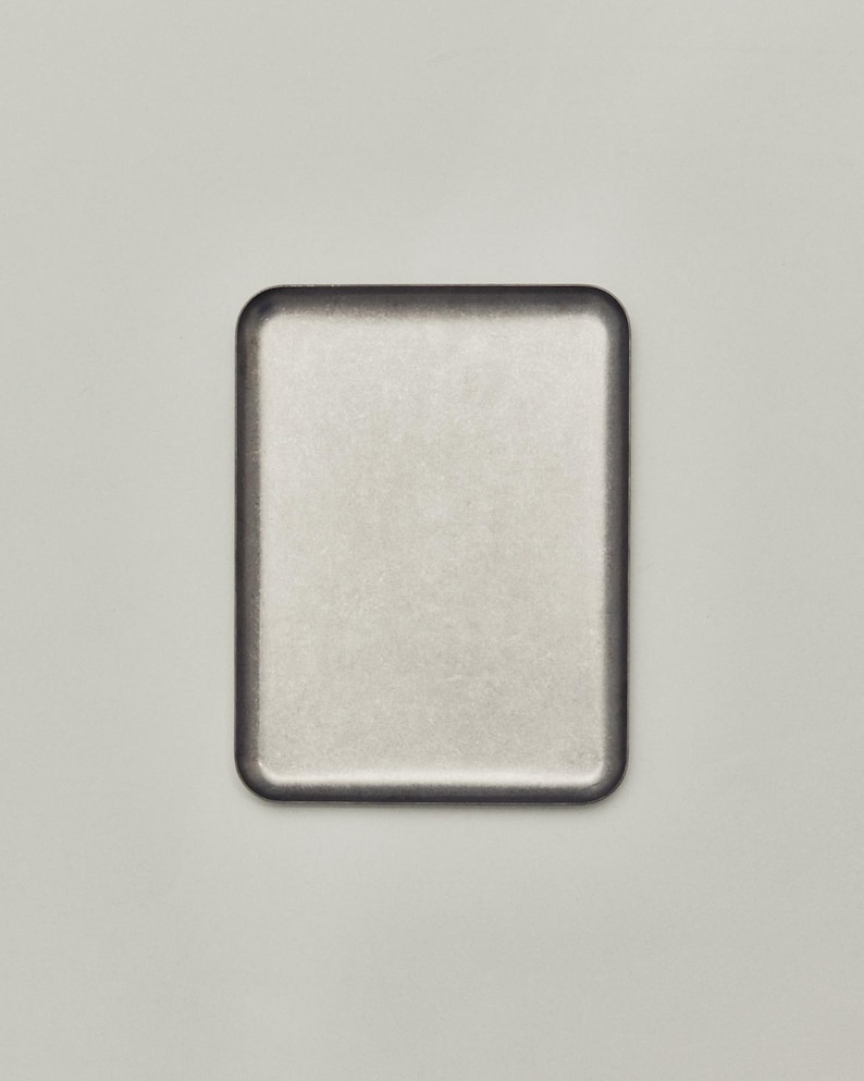 May include: A rectangular, metallic-looking tray with rounded corners. The tray has a light, brushed metal appearance with a darker border. The background is a solid, neutral color.