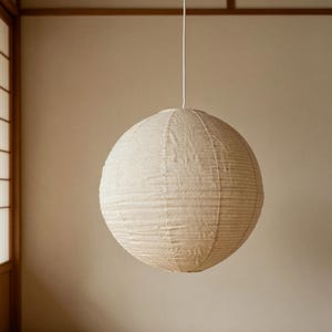 May include: A spherical pendant lamp featuring a textured, off-white paper shade with visible horizontal ribbing. The round light fixture is suspended from a thin, white cord that extends vertically from the top center of the paper sphere.