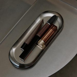 May include: An oval-shaped brushed metal tray containing three cosmetic items: a brown squeeze tube, a black cylindrical lipstick tube labeled Powder Kiss Liquid Lipcolour, and a rectangular lipstick case with a metallic gold cap and amber body.