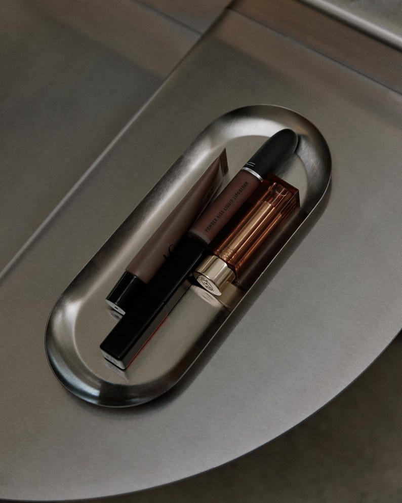 May include: An oval-shaped brushed metal tray containing three cosmetic items: a brown squeeze tube, a black cylindrical lipstick tube labeled Powder Kiss Liquid Lipcolour, and a rectangular lipstick case with a metallic gold cap and amber body.