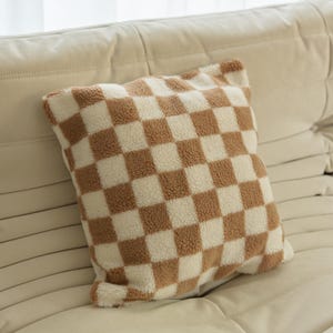 May include: A brown and white checkered throw pillow with a soft, fuzzy texture. The pillow is made of a plush fabric and has a square shape.