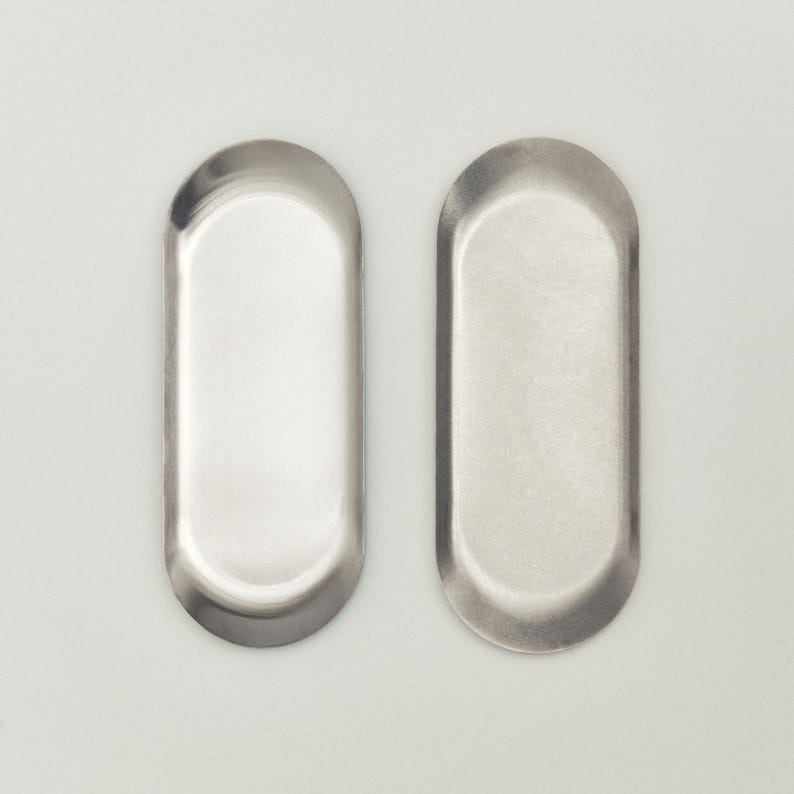 May include: Two oval-shaped metal trays with a smooth, reflective silver finish. Each tray features a shallow basin and raised, rounded edges, presenting a clean, minimalist design suitable for organizing small items on a flat surface.