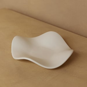 May include: A white ceramic dish with a wavy, organic shape. The dish is sitting on a light brown surface.