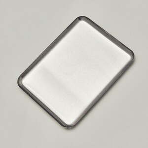 May include: A rectangular, brushed metal tray with rounded corners. The tray has a slightly raised edge and a smooth, reflective surface. The metal has a silver color and a matte finish. The tray is empty.