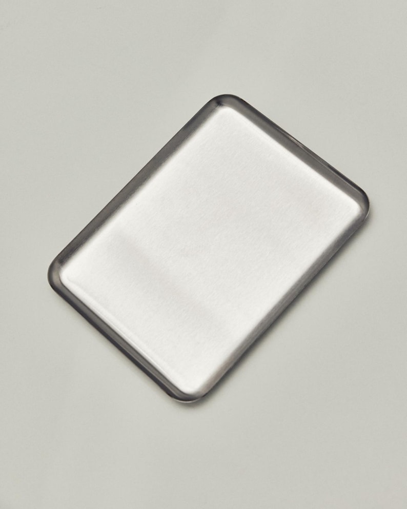 May include: A rectangular, brushed metal tray with rounded corners. The tray has a slightly raised edge and a smooth, reflective surface. The metal has a silver color and a matte finish. The tray is empty.