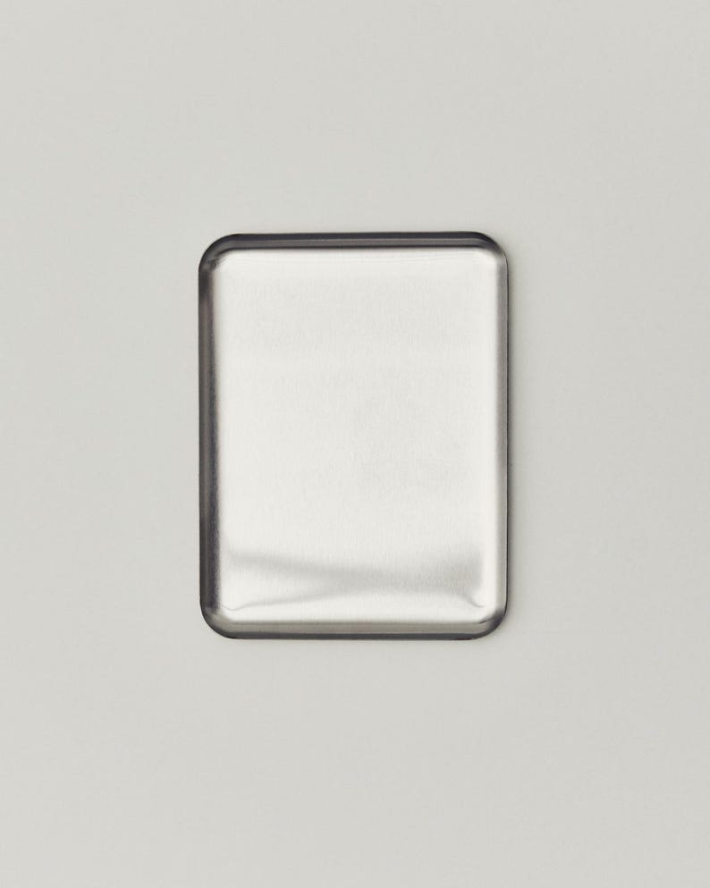 May include: A rectangular, brushed metal object with rounded corners. The metal has a reflective surface, and the edges are outlined with a darker, possibly black, border. The object is set against a plain, light gray background.