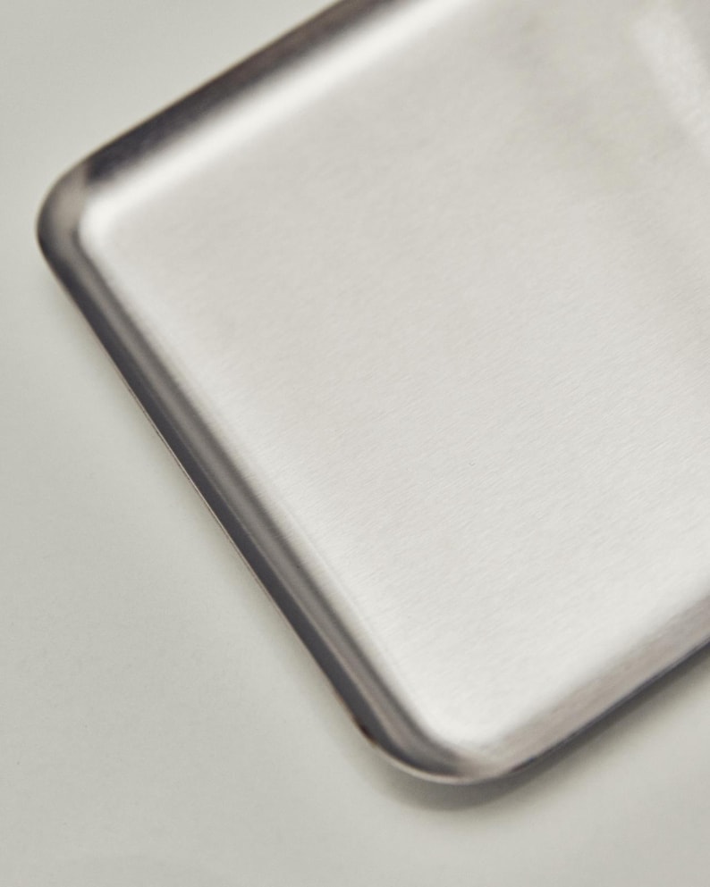 May include: A close-up of a rectangular, brushed metal tray with rounded corners. The tray has a silver finish and a slightly textured surface. The edges are darker than the center, creating a subtle contrast.