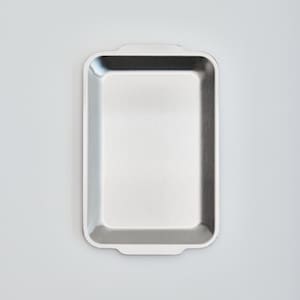 Peut inclure: Un plateau de service rectangulaire en acier inoxydable avec des coins arrondis et de petites poignées. Le plateau a une finition en métal brossé et est vide, sur fond blanc.