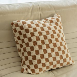 May include: A brown and white checkered throw pillow with a soft, fuzzy texture. The pillow is sitting on a white couch.