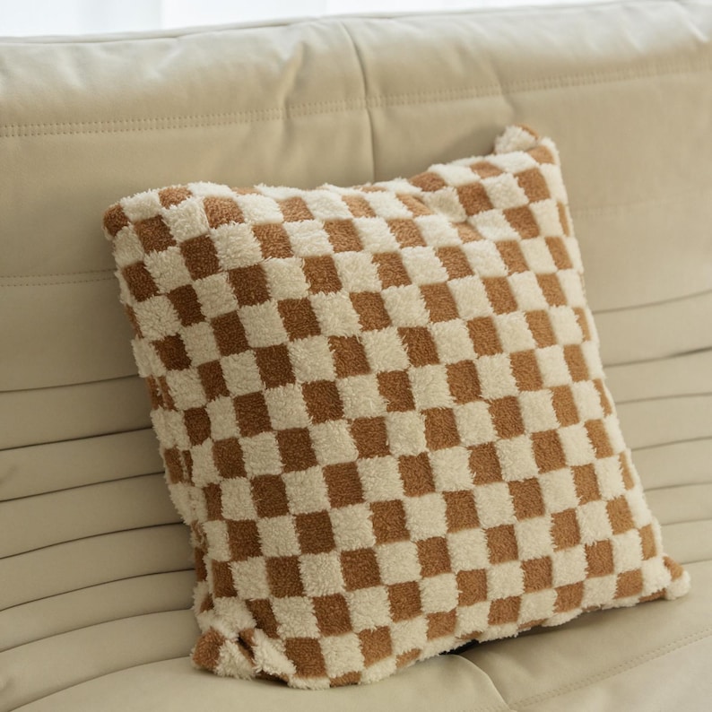 May include: A brown and white checkered throw pillow with a soft, fuzzy texture. The pillow is sitting on a white couch.