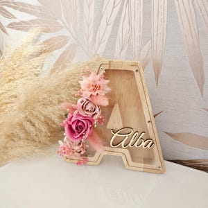 Peut inclure: Boîte à monnaie en bois en forme de lettre A avec un devant transparent et un arrangement floral rose. Le nom "Alba" est écrit sur le devant de la boîte.