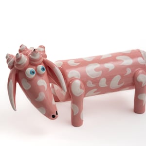 Puede incluir: Escultura de cabra de cerámica rosa con manchas blancas. La cabra tiene grandes ojos azules, cuernos rizados y orejas largas. Un objeto decorativo que añade un toque de alegría a cualquier espacio.