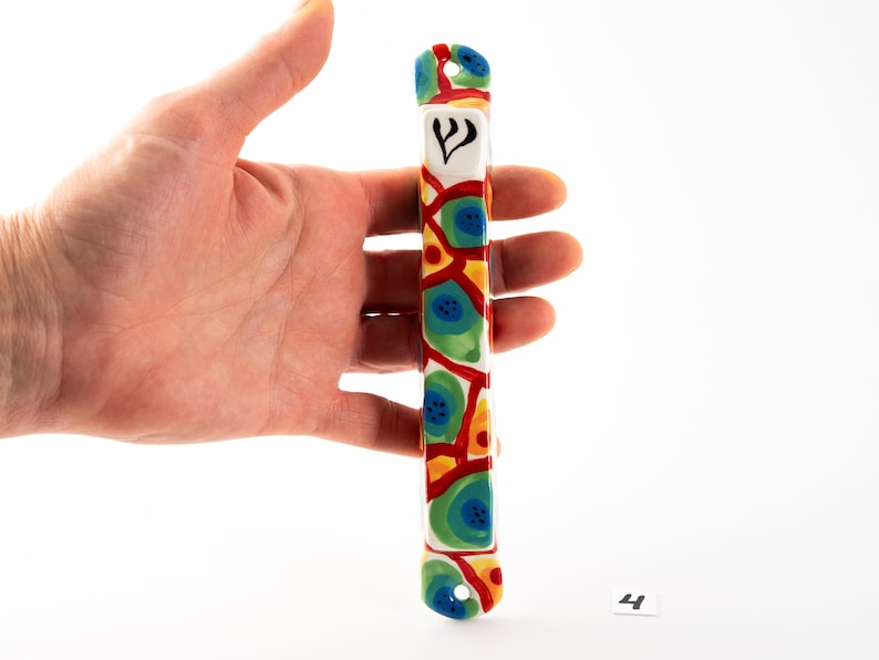 May include: A colorful ceramic mezuzah with a white background and a blue, green, red, and yellow geometric pattern. The mezuzah has a Star of David symbol on the front.